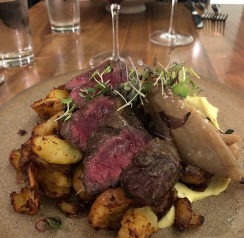 A plate of medium-rare steak slices atop crispy potatoes, garnished with microgreens and accompanied by creamy sauce.