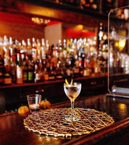 A cocktail glass garnished with lemon twists sits on a woven mat, with a bar and bottles in the background.