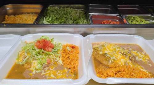 Two takeout containers filled with rice, beans, lettuce, tomatoes, and a cheesy enchilada, with toppings in the background.