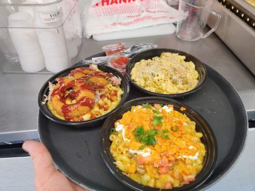 A hand holding a black tray with three bowls of macaroni dishes, each topped with various ingredients and sauces.