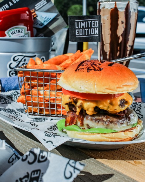 A juicy burger with cheese, lettuce, and tomato, served with sweet potato fries and a milkshake in the background.