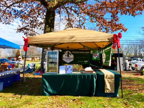 A market stall with a tan canopy, decorated with red bows, showcasing artisanal cheese products under a tree.
