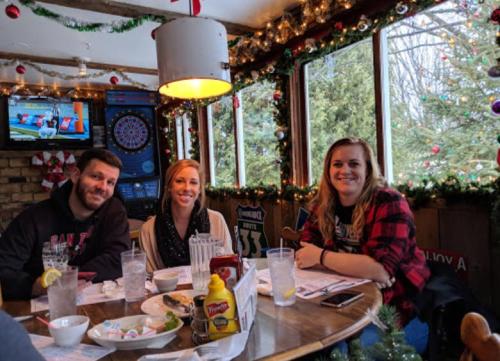 Three friends sit at a table in a festive restaurant, decorated with holiday lights and greenery, enjoying their meal.