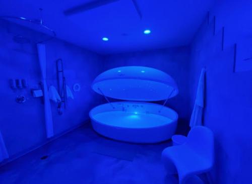 A serene, blue-lit floatation tank in a minimalist room with a chair and soft lighting.