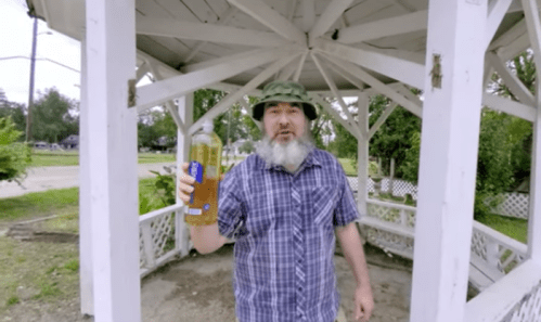A man in a plaid shirt and camouflage hat stands in a gazebo, holding a bottle of drink.