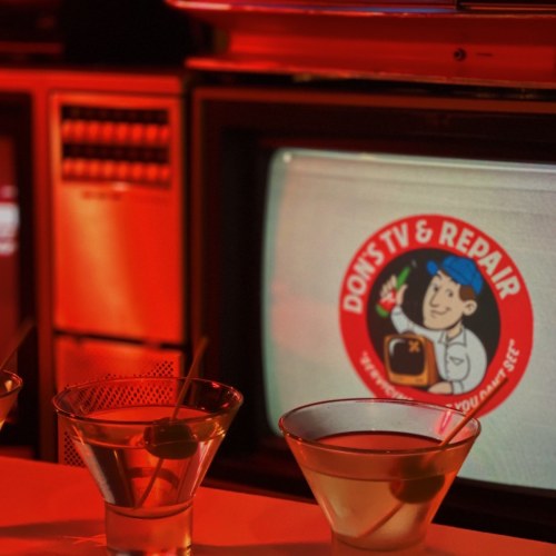 A dimly lit bar scene featuring two martini glasses in front of a vintage TV displaying a repair shop logo.