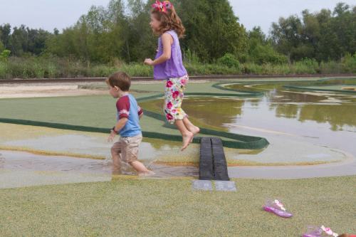 Two children play in a shallow water area at a park, one jumping off a small platform while the other walks through the water.