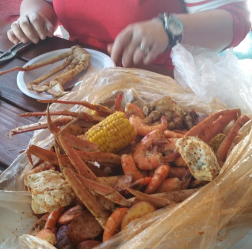 A seafood feast with crabs, shrimp, corn, and potatoes served in a plastic bag on a table. Hands are preparing to eat.