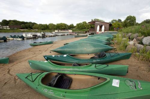 A row of green kayaks lined up on a sandy shore near a calm lake, with a dock and trees in the background.