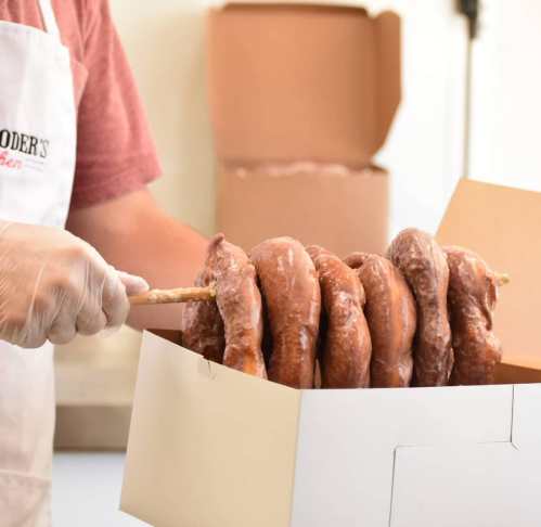 A person in an apron holds a stick with several chocolate-covered donuts over a box.