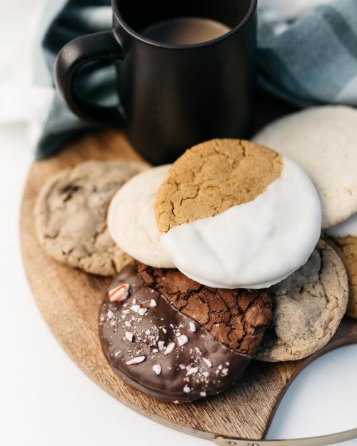 A wooden platter with various cookies and a cup of coffee, featuring chocolate-dipped and frosted cookies.