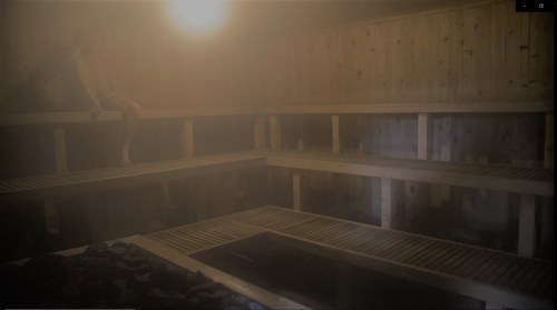 A dimly lit sauna with wooden benches and a person sitting quietly on one of the benches. Steam fills the air.