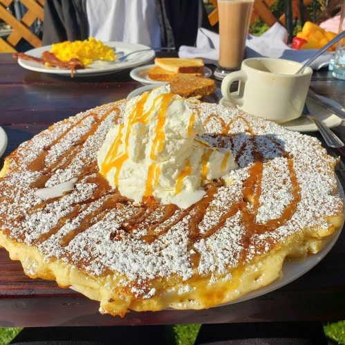 A large, fluffy pancake topped with whipped cream, caramel drizzle, and powdered sugar, served with coffee and breakfast items.