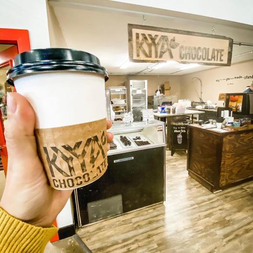 A hand holds a coffee cup in front of a chocolate shop with a wooden sign reading "KYA Chocolate."