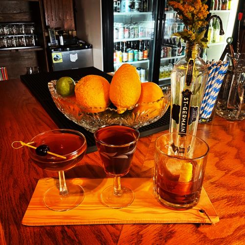 Three cocktails on a wooden board, with a bottle of St-Germain and a bowl of oranges in the background.