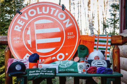 A display of Revolution Bicycles merchandise, including hats and scarves, with a large logo sign in a forest setting.