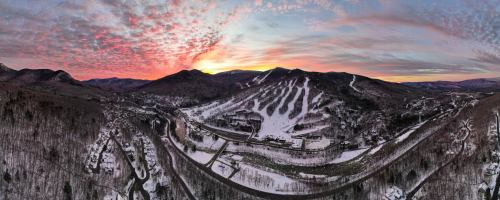 A panoramic view of snow-covered mountains and ski slopes at sunset, with a colorful sky and a nearby village.