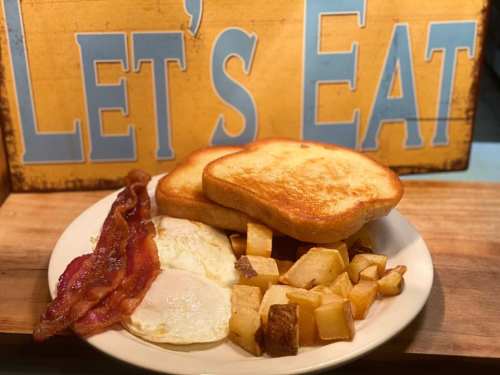 A plate with two fried eggs, crispy bacon, golden toast, and diced potatoes, with a "Let's Eat" sign in the background.