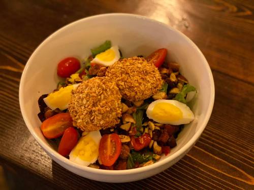 A bowl of salad with greens, cherry tomatoes, boiled eggs, nuts, and two crispy oat-covered patties on top.