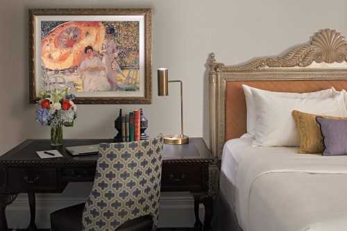 A stylish hotel room featuring a bed, desk with flowers, and a framed painting on the wall.