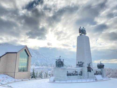 A snowy landscape featuring a large monument with statues, alongside a modern building under a cloudy sky.