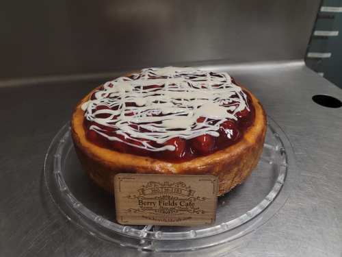 A cheesecake topped with cherry filling and drizzled with white icing, displayed on a clear cake stand.