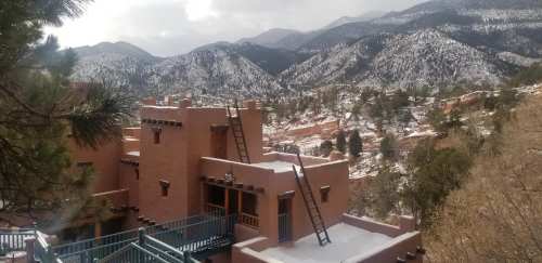 A scenic view of adobe buildings with a ladder, surrounded by snow-capped mountains and pine trees.