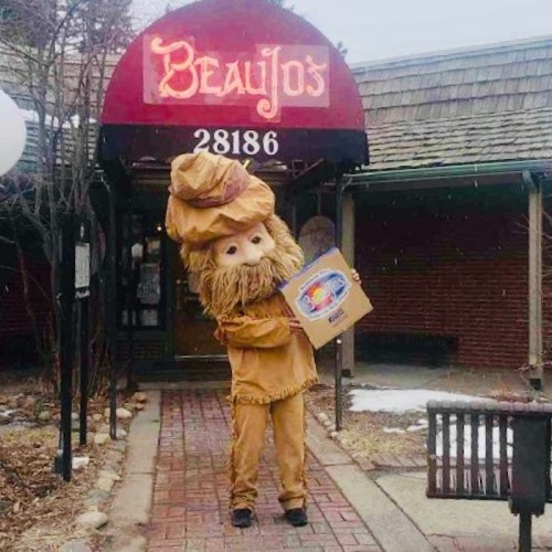 A person in a large mascot costume with a beard and hat stands outside Beau Jo's restaurant, holding a box.