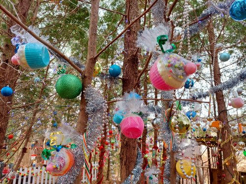 Colorful ornaments and cupcake decorations hang from trees, surrounded by tinsel in a festive outdoor setting.