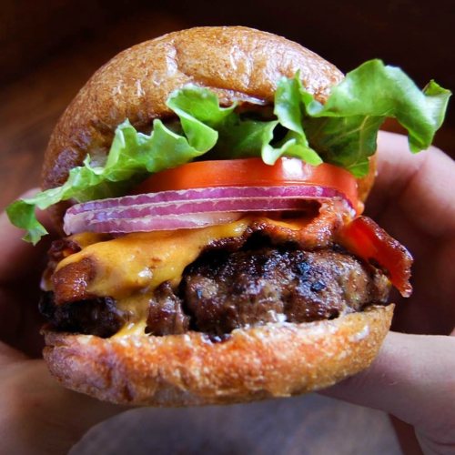A hand holding a juicy burger with lettuce, tomato, onion, bacon, and melted cheese on a toasted bun.