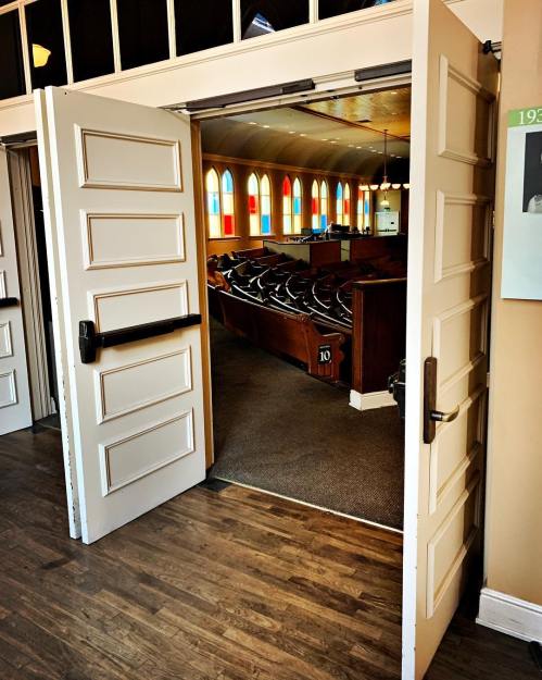 Open double doors leading into a brightly lit church interior with colorful stained glass windows and wooden pews.