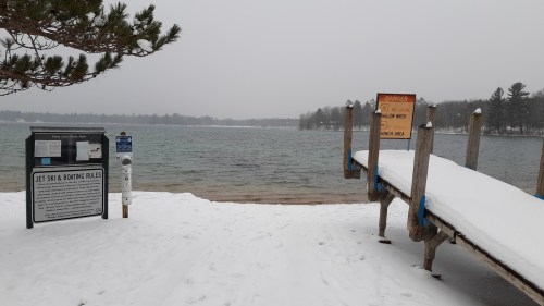 Snow-covered shoreline with a dock, warning sign, and information board by a calm lake on a cloudy day.