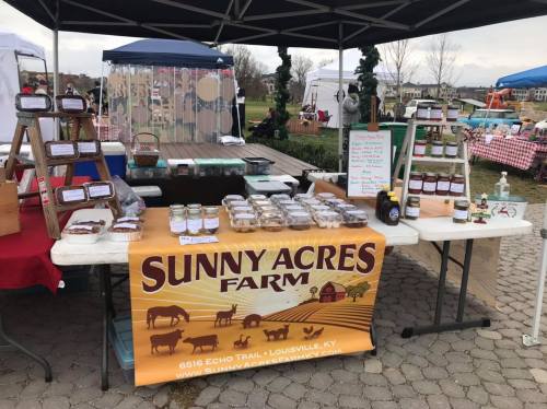 A market stall displaying jars of preserves, baked goods, and a sign for Sunny Acres Farm in Louisville, KY.