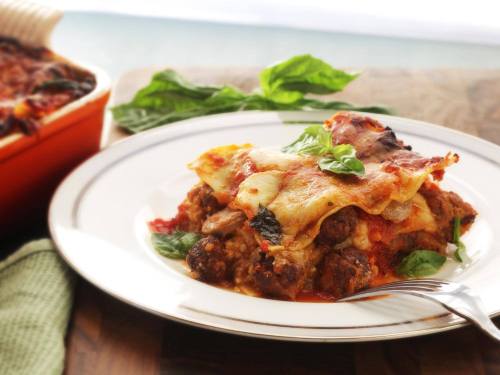 A plate of lasagna topped with fresh basil, served with a fork, beside a baking dish in a bright setting.