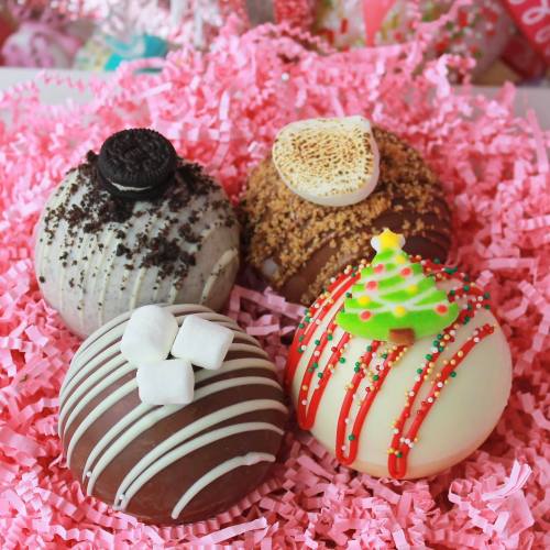 Four decorated chocolate bombs on pink shredded paper: cookies and cream, s'mores, and festive holiday designs.
