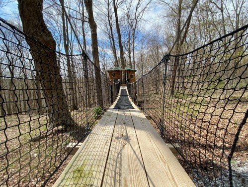 A wooden suspension bridge leads to a small cabin in a wooded area, surrounded by bare trees and gravel.