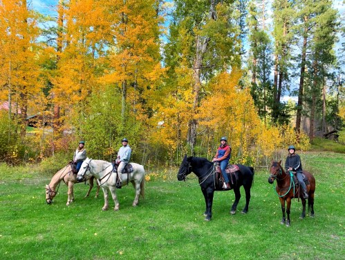 Four riders on horseback in a vibrant autumn setting with colorful trees and green grass.