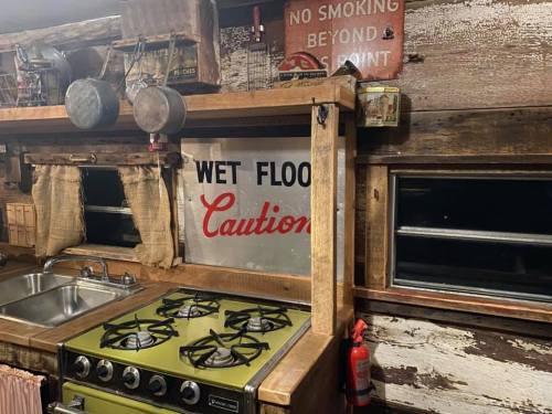 A rustic kitchen with a green stove, metal pots, and a "Wet Floor Caution" sign on the wall.