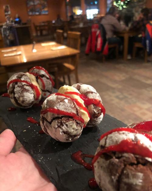 A hand holding a slate with four chocolate dessert balls drizzled with red sauce and topped with cream, in a cozy restaurant.