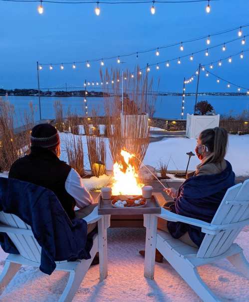 A couple sits by a fire pit on a snowy evening, surrounded by string lights and a scenic waterfront view.