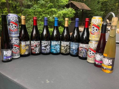 A collection of various beer bottles with colorful labels, arranged on a table in a wooded outdoor setting.
