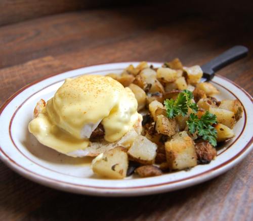 A plate of eggs Benedict topped with hollandaise sauce, served with golden-brown diced potatoes and fresh parsley.