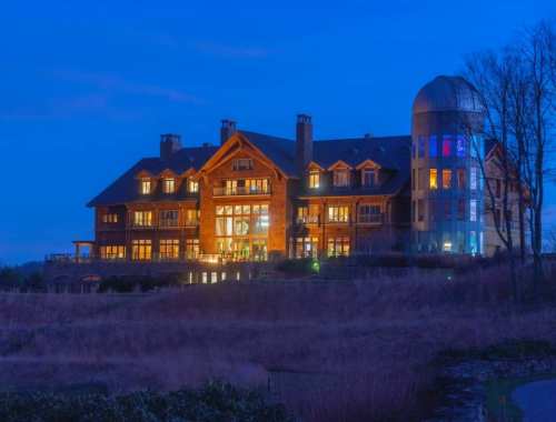 A large, illuminated lodge at dusk, featuring multiple windows and a distinctive tower, surrounded by open fields.