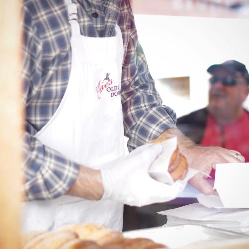 A baker in a white apron hands a pastry to a customer behind a glass display.