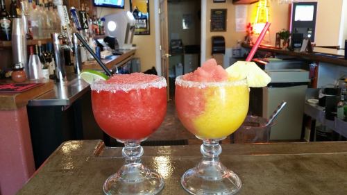 Two colorful frozen cocktails in large glasses, one red with a lime slice and the other yellow with a pineapple wedge.