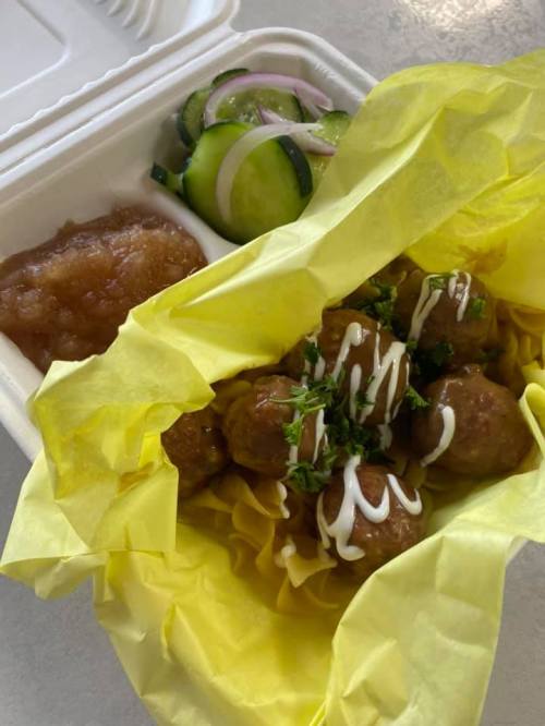 A takeout container with yellow wrapping, featuring meatballs, noodles, cucumber salad, and a side of sauce.