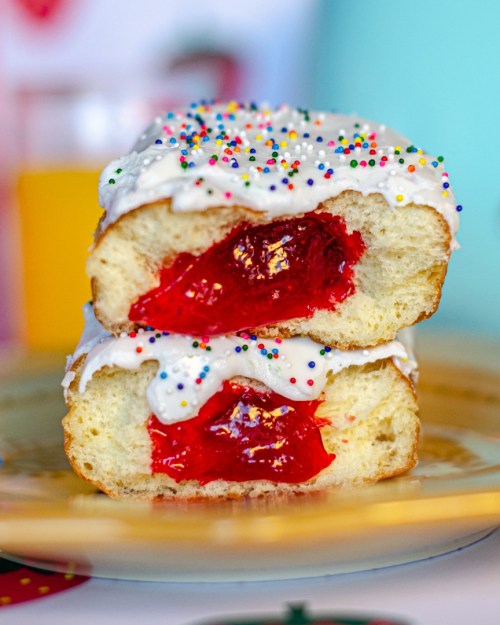 A close-up of a split pastry filled with red jelly, topped with white icing and colorful sprinkles on a gold plate.