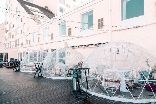 Three transparent dining domes on a wooden deck, surrounded by string lights and a modern building in the background.