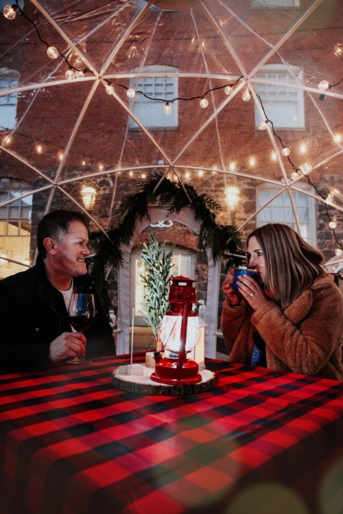 A couple sits inside a cozy, decorated igloo with string lights, enjoying drinks on a checkered tablecloth.