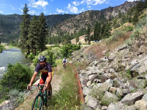 Two cyclists ride along a rocky path beside a river, surrounded by trees and mountains under a blue sky.